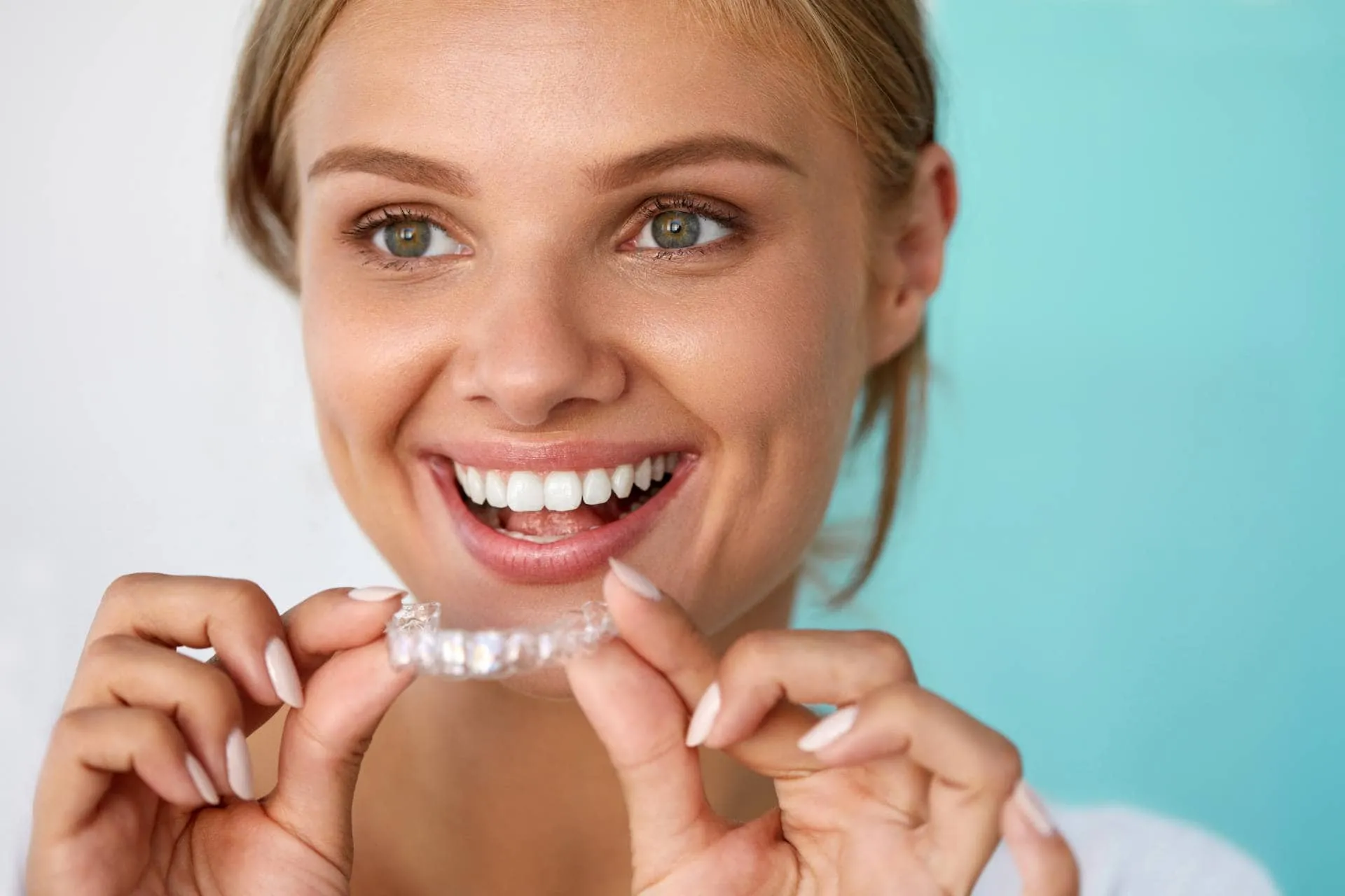 Smiling woman holding clear Invisalign aligner against a light blue background, representing discreet orthodontic treatment for adults.