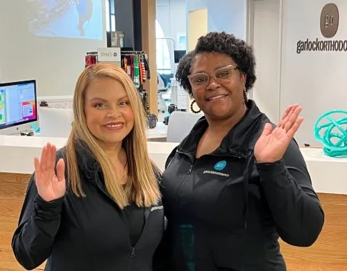 Two friendly staff members at Garlock Orthodontics smiling and waving in the office, showcasing a welcoming atmosphere for patients seeking orthodontic care.