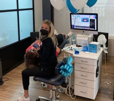 Orthodontist assisting a patient in a modern office setting, featuring dental equipment, balloons, and a computer display, emphasizing patient comfort and care at Garlock Orthodontics.